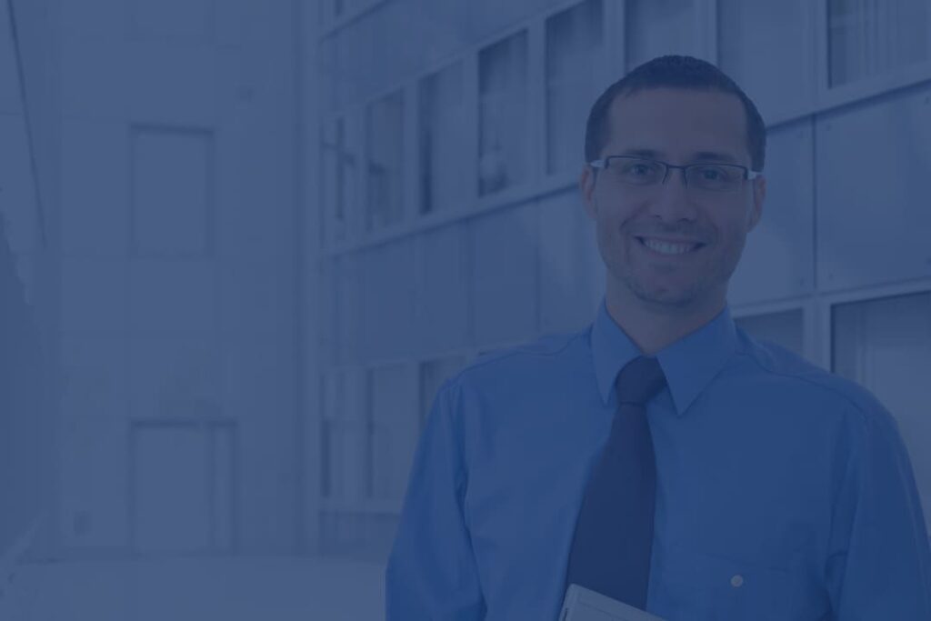 Businessman wearing glasses, a blue shirt, and a tie, standing confidently in front of a modern office building.