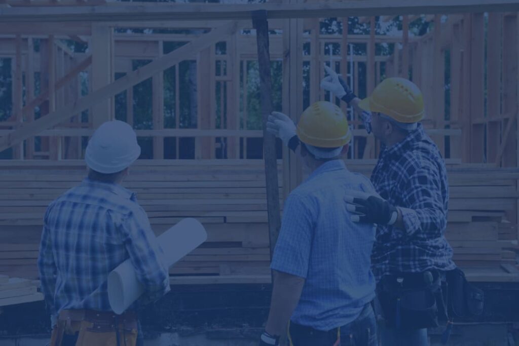 Construction team wearing hard hats and safety gear reviewing blueprints and discussing progress at a wooden house framing site.