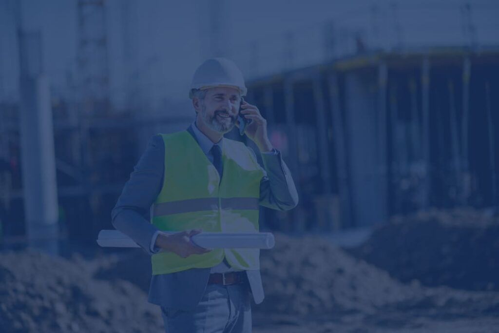 Construction project manager wearing a hard hat and high-visibility vest, holding blueprints while talking on the phone at an active building site.