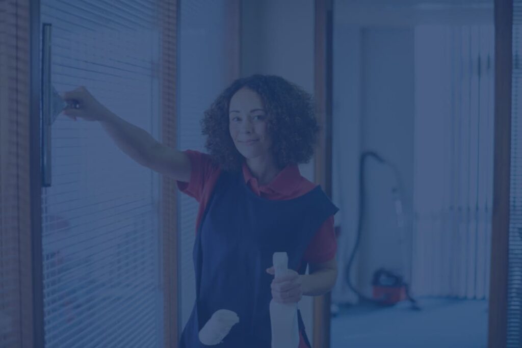 Smiling professional cleaner in a uniform wiping a glass window with a squeegee while holding a spray bottle in an office setting.